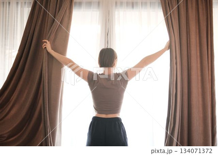 A woman spreads the curtains wide, letting bright daylight enter the room. The scene conveys a new beginning, energy, and a refreshing morning 134071372