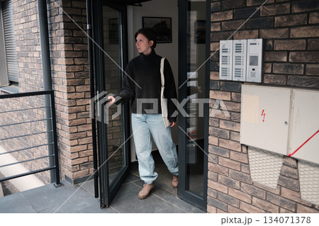 A woman walks out of a building holding the door, wearing a warm sweater and jeans. The scene reflects everyday city movement and lifestyle habits 134071378