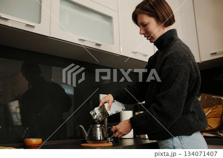 A woman pours loose tea leaves into a glass teapot, preparing a warm drink in a cozy kitchen setting 134071407