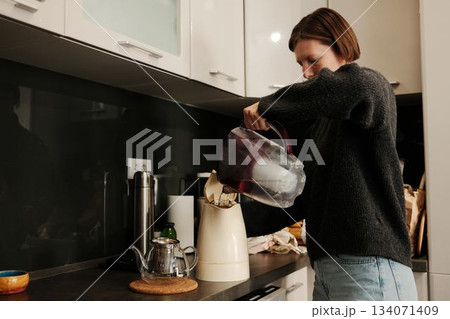 A woman pours filtered water into an electric kettle in a calm kitchen environment, beginning a simple tea-making routine 134071409