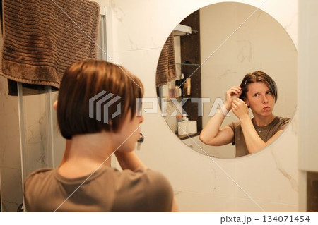 A woman uses a hairpin while looking into a mirror, adjusting her hairstyle as part of her organized morning grooming routine A woman uses a hairpin while looking into a mirror, adjusting her hairstyle as part of her organized morning grooming routine 134071454