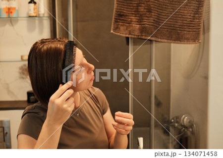 A woman carefully combs her hair in a bathroom, creating a calm and intimate moment of personal care at the start of the day A woman carefully combs her hair in a bathroom, creating a calm and intimate moment of personal care at the start of the day 134071458