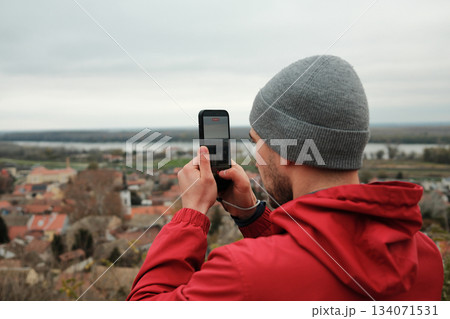 Man in grey beanie and red jacket recording city scenery with a smartphone. The frame reflects travel, technology, and personal experience. Travel blogger concept 134071531