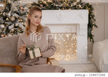 Caucasian woman holding gift box in cozy Christmas room with tree and white fireplace Caucasian woman holding gift box in cozy Christmas room with tree and white fireplace 134071560