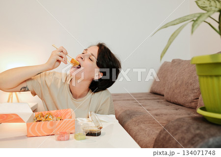 A woman leans toward the table and takes a large bite of fried sushi with chopsticks. The scene emphasizes appetite, strong flavor, and casual dining at home 134071748