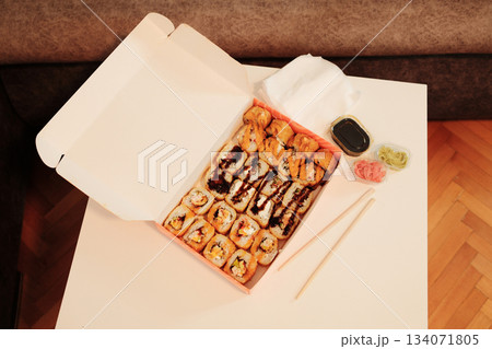An open delivery box with assorted fried sushi rolls, soy sauce, ginger, wasabi, and chopsticks on a white table. A cozy interior setting highlights modern takeaway dining and Asian food culture An open delivery box with assorted fried sushi rolls, soy sauce, ginger, wasabi, and chopsticks on a white table. A cozy interior setting highlights modern takeaway dining and Asian food culture 134071805
