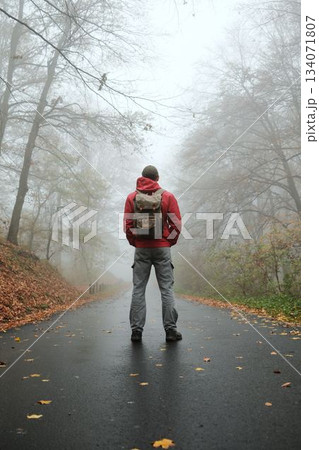 Man with backpack stands still on wet autumn road surrounded by fog and fallen leaves. Back rear view. Quiet and cinematic scene Man with backpack stands still on wet autumn road surrounded by fog and fallen leaves. Back rear view. Quiet and cinematic scene 134071807