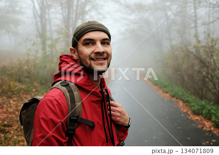 Happy male traveler in red jacket smiling on quiet misty forest road during autumn walk. Solo travel concept 134071809
