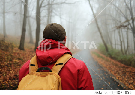 Back rear view of male traveler in red jacket and backpack looking at foggy autumn forest road 134071859