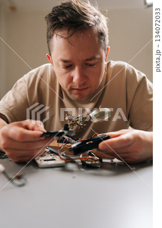 Vertical portrait of serious focused technician examining complex circuit board with magnifying glass, performing detailed electronic repair work, problem-solving, and developing new technology. 134072033