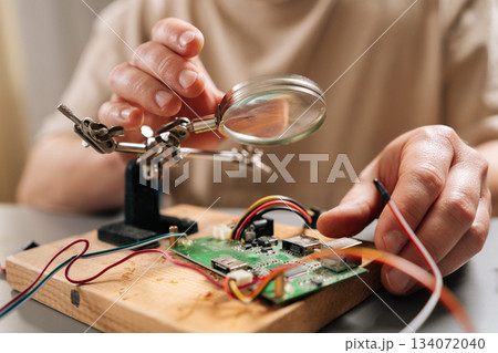 Close-up hands of technician focusing on intricate details of circuit board on wooden base with magnifying glass, using helping hands tools and wires during electronic repair or assembly process. 134072040