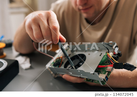 Close-up cropped shot of technician working on electronic device, holding computer hardware with exposed cooling fan, performing repairs and maintenance on tech equipment in modern workshop. Close-up cropped shot of technician working on electronic device, holding computer hardware with exposed cooling fan, performing repairs and maintenance on tech equipment in modern workshop. 134072045
