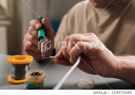 Close-up cropped shot of person soldering wires using electric soldering iron, creating smoke, repairing damaged electrical cable, performing electronic circuit work in modern workshop environment. 134072048