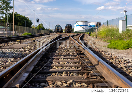 Rusty railway switch toward parked tank wagons under bright blue sky background sunny day. Industrial yard shows metal tracks gravel storage tanks oil refinery plant factory station 134072541