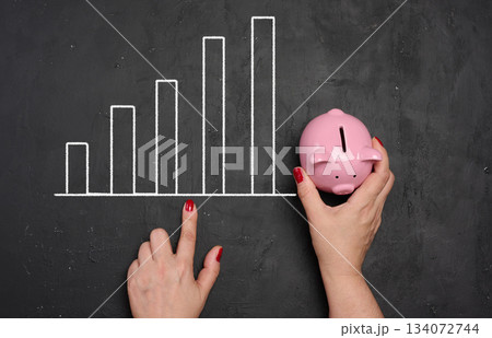 Woman's hand holds a pink ceramic piggy bank against a chalkboard background with a growing graph Woman's hand holds a pink ceramic piggy bank against a chalkboard background with a growing graph 134072744