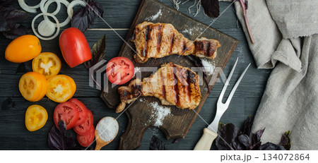 Grilled pork steaks on a wooden board, surrounded by vegetables and basil sprigs. Black table Grilled pork steaks on a wooden board, surrounded by vegetables and basil sprigs. Black table 134072864