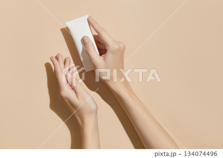 A womans hands holding a white cosmetic tube on a beige background. Beauty treatment concept A womans hands holding a white cosmetic tube on a beige background. Beauty treatment concept 134074496