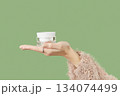 A womans hand in a sweater holds a jar of cream on a green background. Self care beauty treatment 134074499