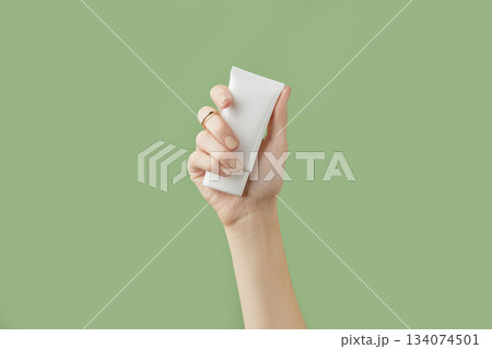 A womans hand holding a white cosmetic tube on a green background 134074501