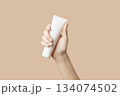 A womans hand holding a white cosmetic tube on a beige background. Care products 134074502