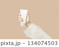 A womans hand in a sweater holds a tube of cream on a beige background. Body care beauty treatment 134074503