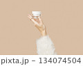 A womans hand in a sweater holds a jar of cream on a beige background. Body care beauty treatment 134074504