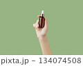 A womans hand holding a brown bottle of serum on a green background 134074508