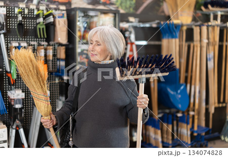 Senior woman in hardware store examines broom and besom. 134074828