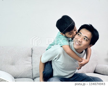 Asian father smiles as his son hugs and kisses him from behind on the couch in a joyful home moment 134074943