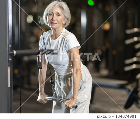 Senior woman performing triceps pulldown on cable machine in gym Senior woman performing triceps pulldown on cable machine in gym 134074981