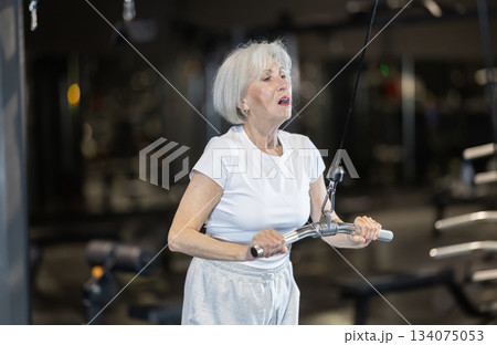 Senior woman performing triceps pulldown on cable machine in gym 134075053