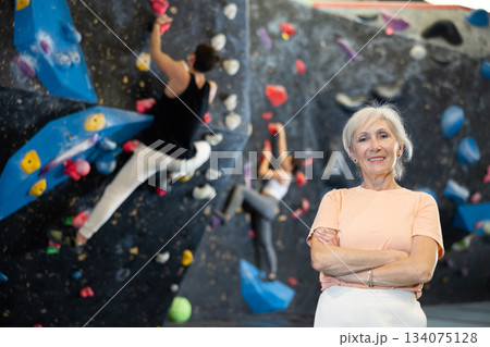 Elderly woman in sportswear smiling at camera while standing against artificial training climbing rock wall in adventure park 134075128
