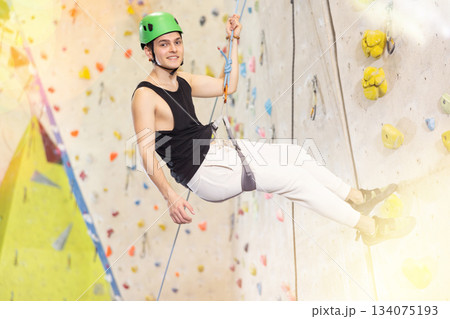 Well-equipped, positive young man going down on rope after reaching top in indoor bouldering adventure park Well-equipped, positive young man going down on rope after reaching top in indoor bouldering adventure park 134075193