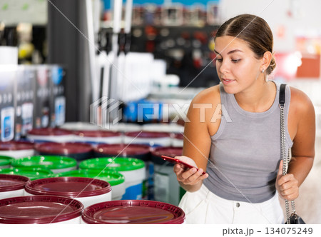 Woman examine bucket of paint, buy, collect necessary supplies in store according to list for repairs Woman examine bucket of paint, buy, collect necessary supplies in store according to list for repairs 134075249