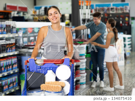 Woman shopper stand with trolley filled with paint goods for repair and coloring in store sales hall. Woman shopper stand with trolley filled with paint goods for repair and coloring in store sales hall. 134075410