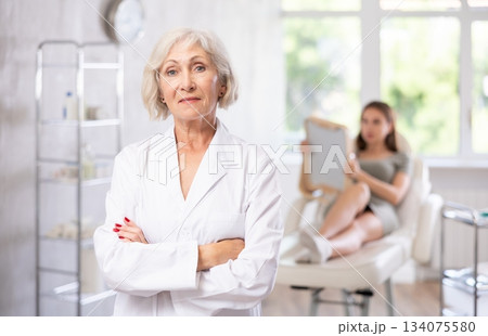 Smiling senior woman doctor in throws up his hands with woman patient looking at mirror behind in clinic. Smiling senior woman doctor in throws up his hands with woman patient looking at mirror behind in clinic. 134075580