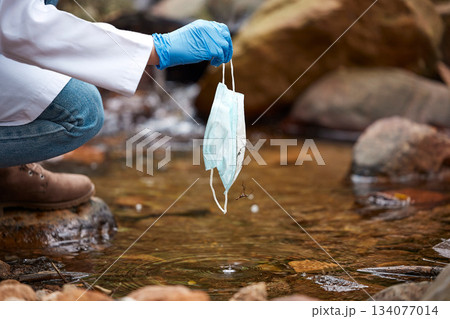 Pollution, litter and face mask in river with hand of person for nature conservation, environment and safety. Covid, danger and trash with garbage in water for medical waste, healthcare and cleaning 134077014