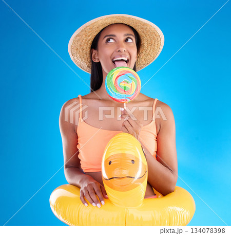 Pool float, happy woman and lollipop lick in a studio with sweet, swimsuit and sweets. Isolated, blue background and holiday of a young female with happiness, licking candy and feeling funny or silly 134078398