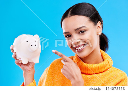 Happy woman, money and pointing to piggy bank for investment, budget or finance against blue studio background. Portrait of female with savings, cash or coin for profit investing or financial freedom Happy woman, money and pointing to piggy bank for investment, budget or finance against blue studio background. Portrait of female with savings, cash or coin for profit investing or financial freedom 134078558