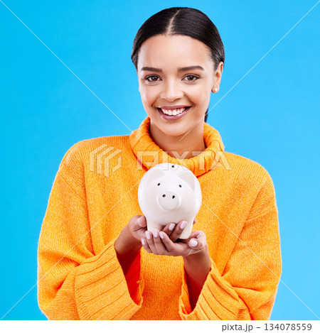 Happy woman, money and piggy bank for investment, budget or finance against a blue studio background. Portrait of female smile holding savings, cash or coin for profit, investing or financial freedom 134078559