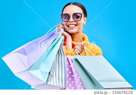 Retail bags, studio and woman portrait with a smile and happiness from boutique sale. Happy, shopping and female model with store bag and sales choice in isolated blue background with sunglasses Retail bags, studio and woman portrait with a smile and happiness from boutique sale. Happy, shopping and female model with store bag and sales choice in isolated blue background with sunglasses 134079129