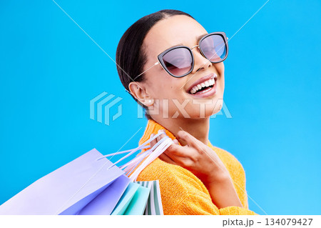 Shopping bags, retail and woman portrait with a smile and sunglasses from boutique sale. Happy, customer and female model with store bag and sales choice in isolated blue background in studio Shopping bags, retail and woman portrait with a smile and sunglasses from boutique sale. Happy, customer and female model with store bag and sales choice in isolated blue background in studio 134079427