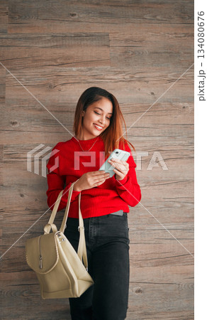 Phone, fashion and a woman in studio on a wooden background typing a text message or reply. Mobile, contact and attractive young female browsing social media while carrying a handbag in red clothes Phone, fashion and a woman in studio on a wooden background typing a text message or reply. Mobile, contact and attractive young female browsing social media while carrying a handbag in red clothes 134080676