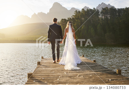 Love, wedding and back of couple by a lake standing, bonding and holding hands on the pier. Nature, romance and young bride and groom in an intimate moment together outdoor on a romantic marriage day Love, wedding and back of couple by a lake standing, bonding and holding hands on the pier. Nature, romance and young bride and groom in an intimate moment together outdoor on a romantic marriage day 134080733