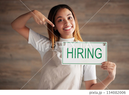 Happy woman, portrait and pointing to hiring sign for small business recruitment, career or job opportunity against studio background. Female entrepreneur with apron holding billboard poster for hire Happy woman, portrait and pointing to hiring sign for small business recruitment, career or job opportunity against studio background. Female entrepreneur with apron holding billboard poster for hire 134082006