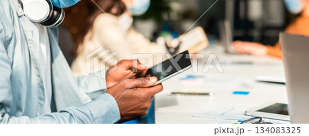 Education And Work During Pandemic. Closeup of african american man wearing surgical face mask and using digital tablet, sitting at desk. Group of diverse people sitting at table in blurred background 134083325