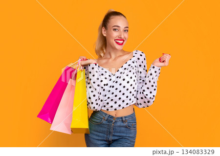 Fashion And Style. Positive Young Woman Holding Colorful Shopping Bags With Gifts And Purchases, Standing And Posing At Studio Isolated Over Pastel Orange Background Wall, Banner, Copy Space 134083329