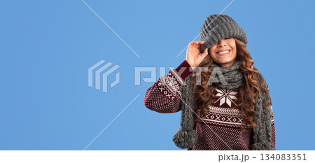 Winter fashion. Young girl pulling down warm beanie hat and smiling over blue background. Panorama 134083351
