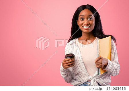 Smiling African American woman with notebooks and takeaway coffee on pink studio background. Portrait of black female student looking at camera, ready to start school. Education concept 134083416