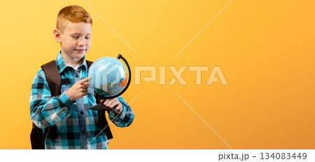 Positive schoolboy with backpack holding globe, studying geography on yellow studio background, copy space. Happy boy going to school, enjoying his school days. Kids education concept 134083449